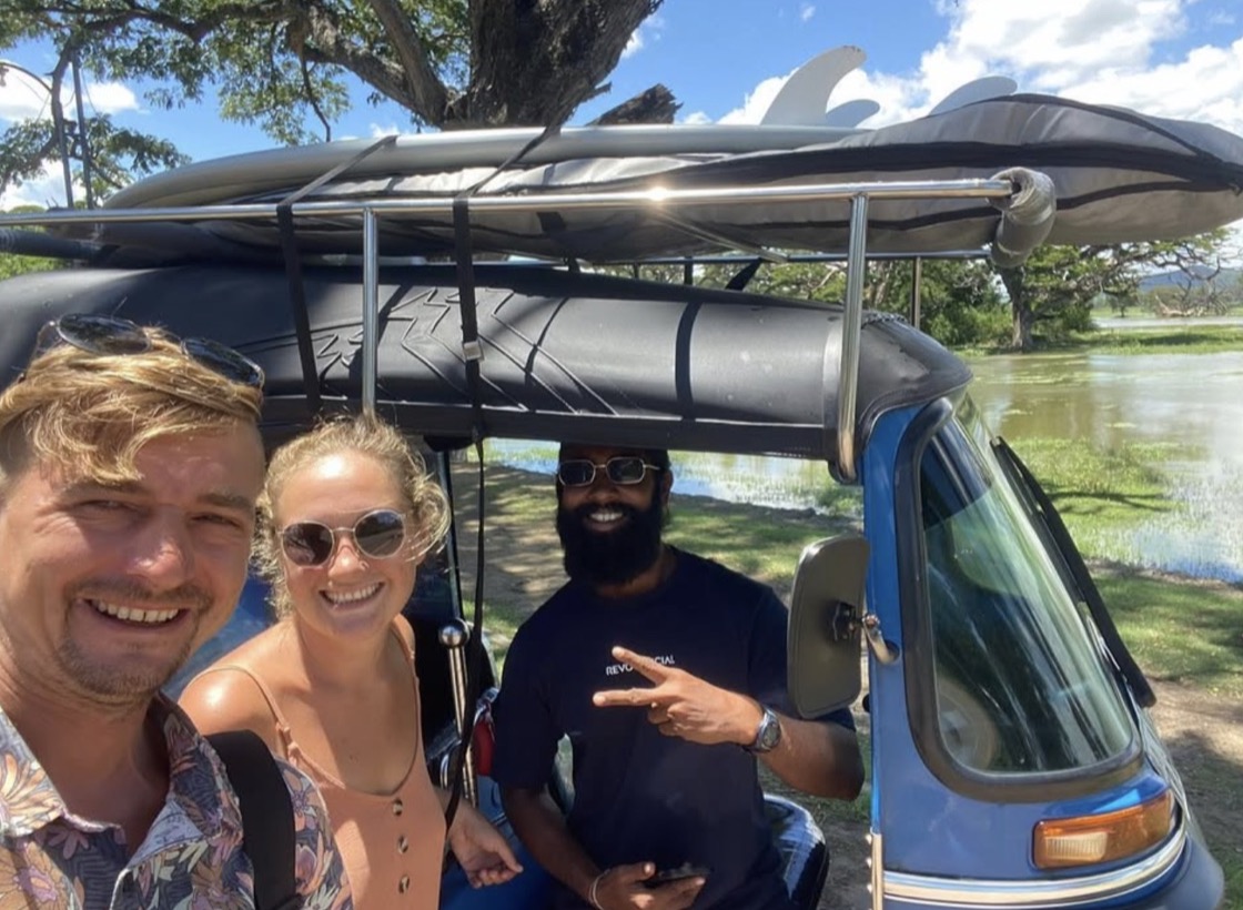 Roma, Maria and Lahiru smiling beside a blue tuk-tuk carrying surfboards